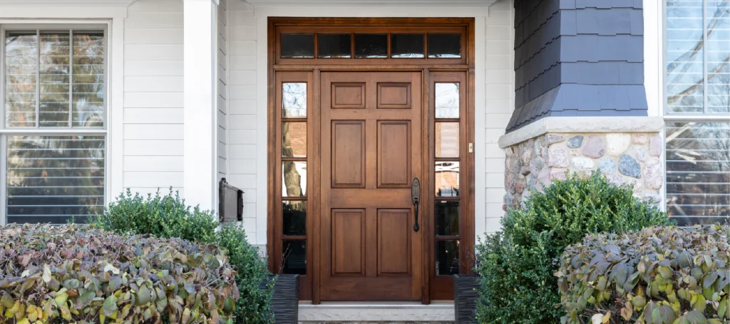 The front entrance of a house features a dark wooden door with glass panels, flanked by two large windows and surrounded by green shrubs